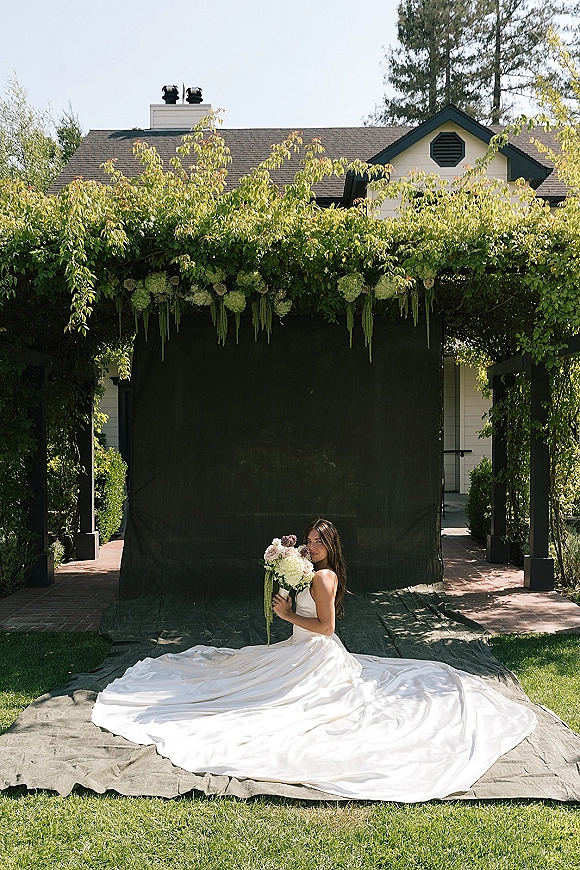 Bridal portrait of a bride seated on her long train holding a white and mauve bouquet beneath a floral arch on a garden lawn with brick walkway