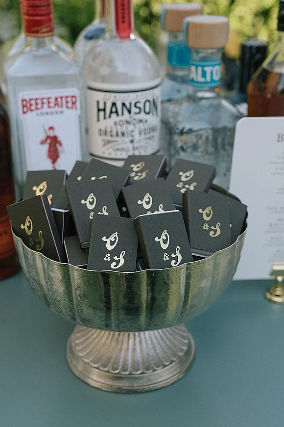 Matchbook wedding favors displayed in a metal pedestal bowl, personalized matchbooks with monogram printing beside liquor bottles on a bar tabletop
