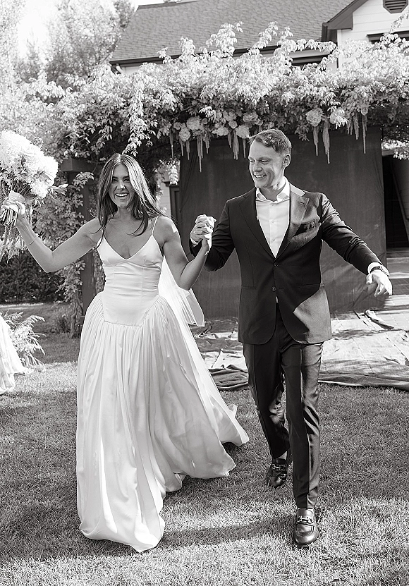 Wedding recessional as bride and groom walking hand in hand, bride holding bouquet beneath a floral arch on an outdoor garden lawn