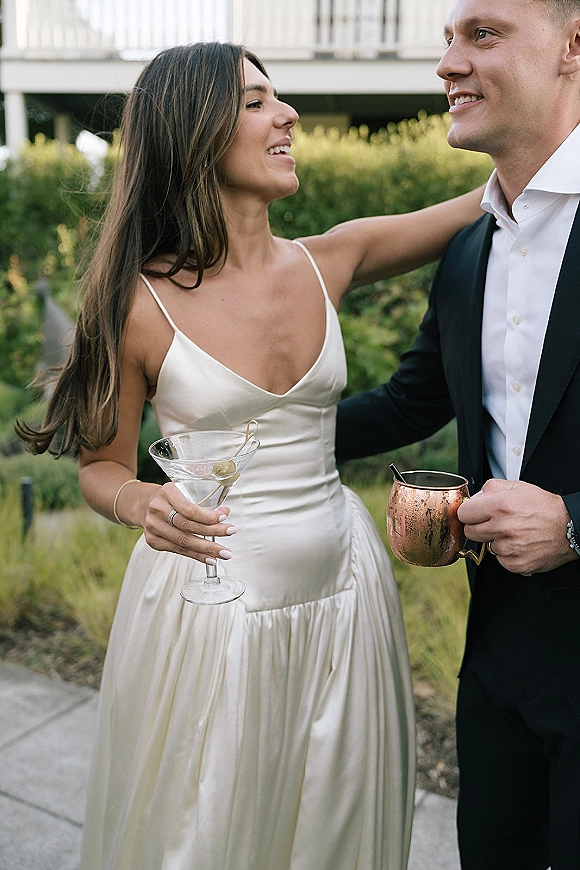 Couple portrait of bride and groom candid laughing arm in arm, holding martini and copper mug on a garden walkway with white railing