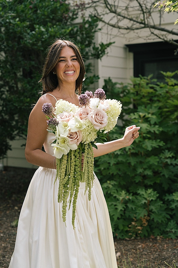 Bridal portrait of a smiling bride holding bouquet with blush roses and orchids, wearing a spaghetti strap gown in greenery by a house window