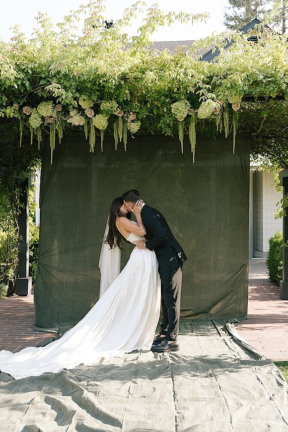 Wedding kiss portrait of bride and groom kissing under a garden pergola with a veil accent, hydrangea garland, and draped backdrop