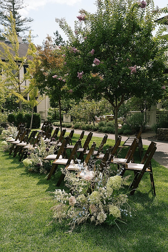 Outdoor ceremony setup with wood folding chairs and ceremony aisle flowers, lined with white and green roses on a garden lawn walkway