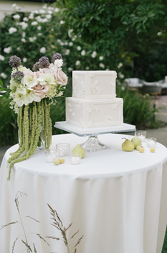 Wedding cake two-tier square design with white embossed floral frosting on a stand, surrounded by roses, orchids, fruit, and candles in a garden setting