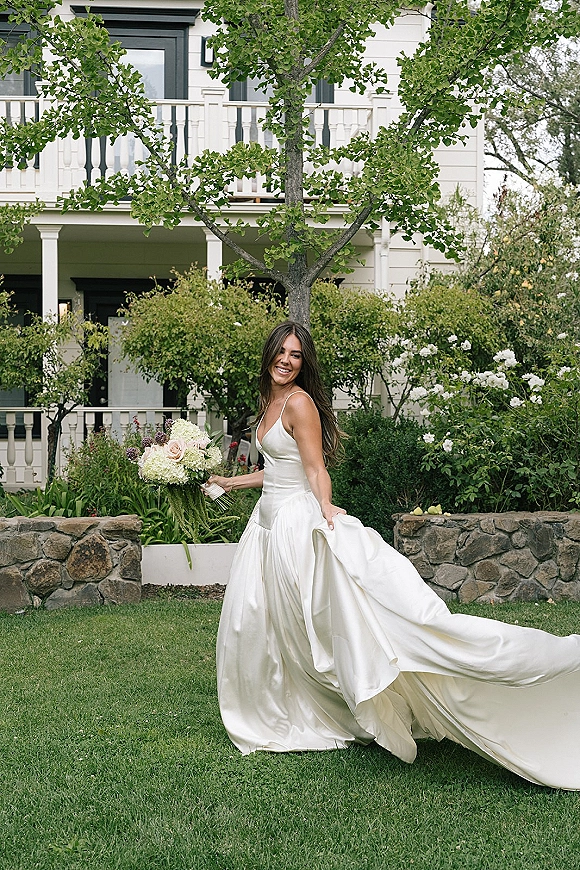Bridal portrait of a bride in a wedding dress with a long train, holding a white and blush bouquet on a garden lawn by a stone wall