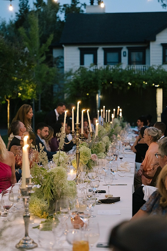 Reception tablescape with a long banquet table wedding setup, white linens, silver candelabras, and taper candles under string lights at dusk