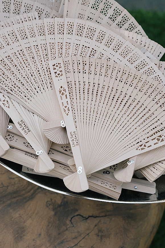 Wedding hand fans with personalized text and laser-cut wood design, fastened with metal rivets, arranged in a metal bowl on a wooden table