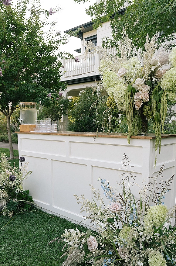 Wedding bar setup with white outdoor wedding bar, hydrangeas and roses, hanging amaranthus, glassware, and beverage dispenser on a lawn by a white house porch
