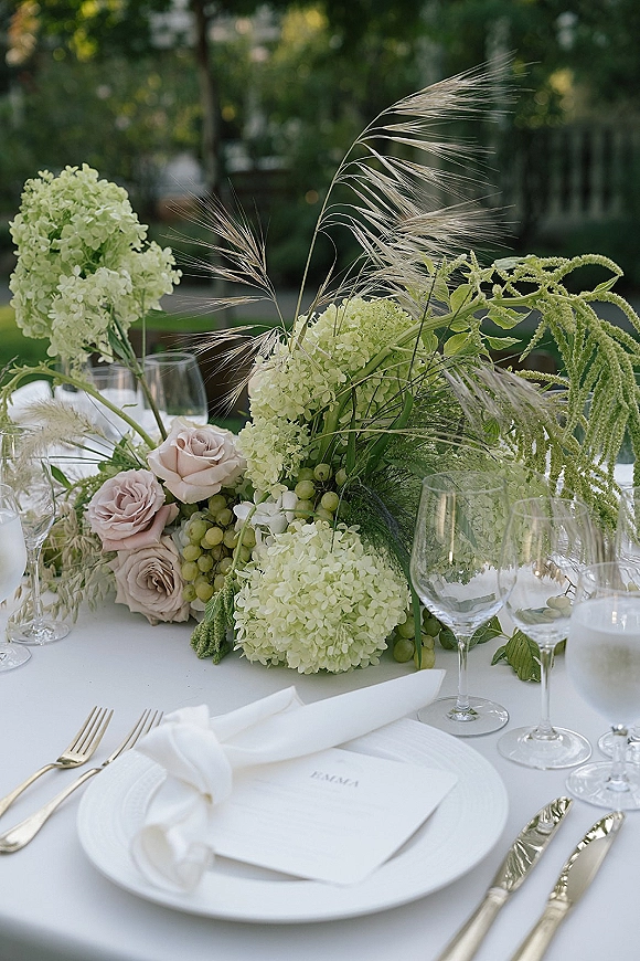 Reception tablescape with wedding table centerpiece of green hydrangeas and blush roses, gold flatware on white linens in a garden setting