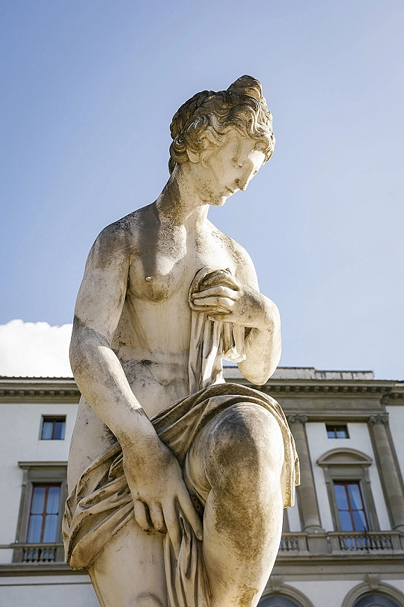 Stone garden statue, classical marble statue with draped fabric carving set before an ornate building facade and blue sky with clouds