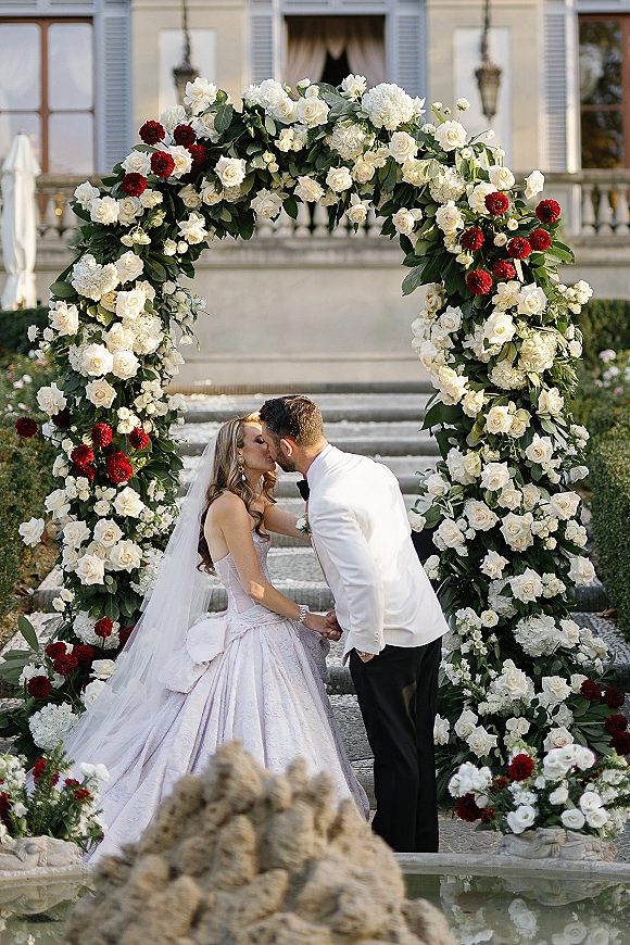 Wedding kiss portrait of bride and groom kissing beneath a white rose floral arch, her long veil flowing on stone steps by a mansion facade