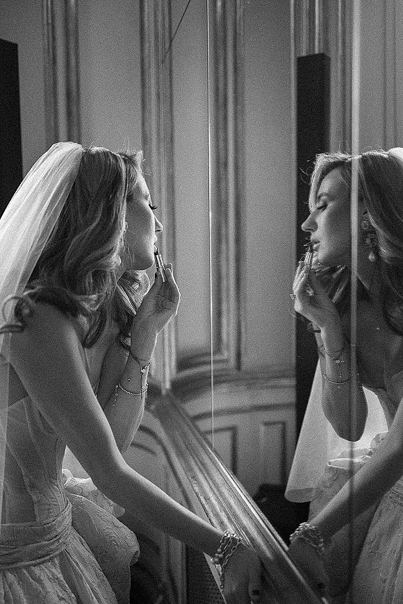 Bridal getting ready, bride applying lipstick in a mirror while wearing a strapless lace wedding dress beside draped curtains and a wood banister