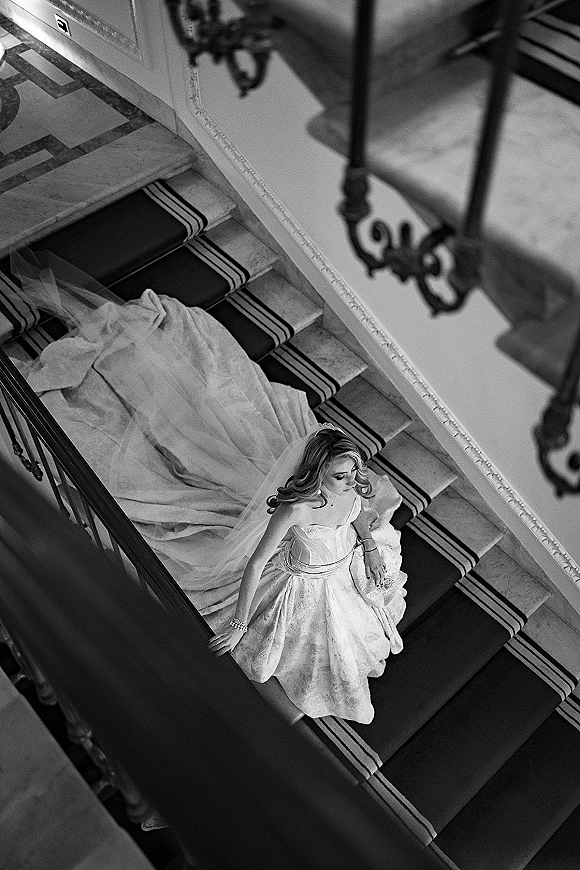 Bridal portrait of a bride on staircase, seated in a strapless gown with long train and veil cascading down the stairs, looking down