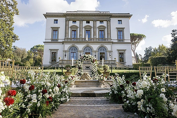 Ceremony setup for an outdoor wedding ceremony with floral arch, red and white aisle flowers, and gold Chiavari chairs on a villa terrace