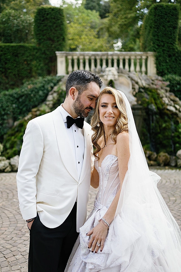 Couple portrait of bride and groom portrait with groom whispering as she looks at camera, veil and lace dress by stone balustrade in garden courtyard
