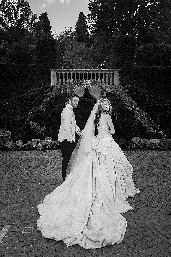 Couple portrait in a black and white wedding portrait, holding hands as they look back by a garden fountain and cobblestone courtyard