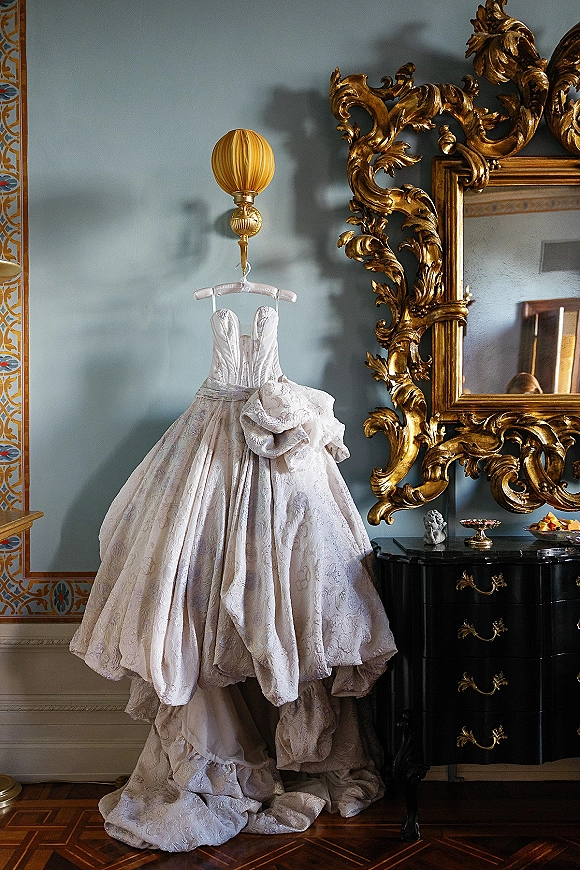 Wedding dress strapless ball gown hanging on a dresser beneath an ornate gold mirror, jacquard fabric and draped overskirt against a blue wall