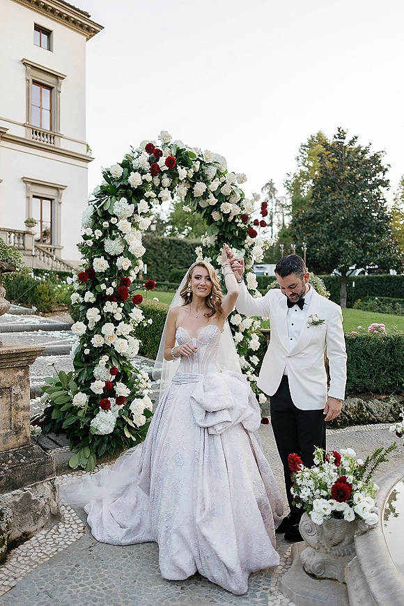 Newlywed portrait of bride and groom under a white rose floral arch, holding hands by a stone fountain with villa garden hedges behind