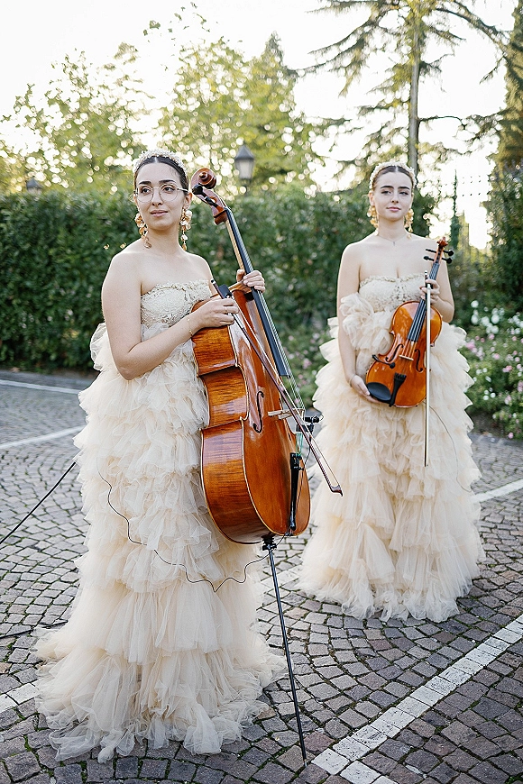 Wedding musicians perform as a string duo wedding, cellist and violinist in matching tulle gowns playing in a garden on cobblestones