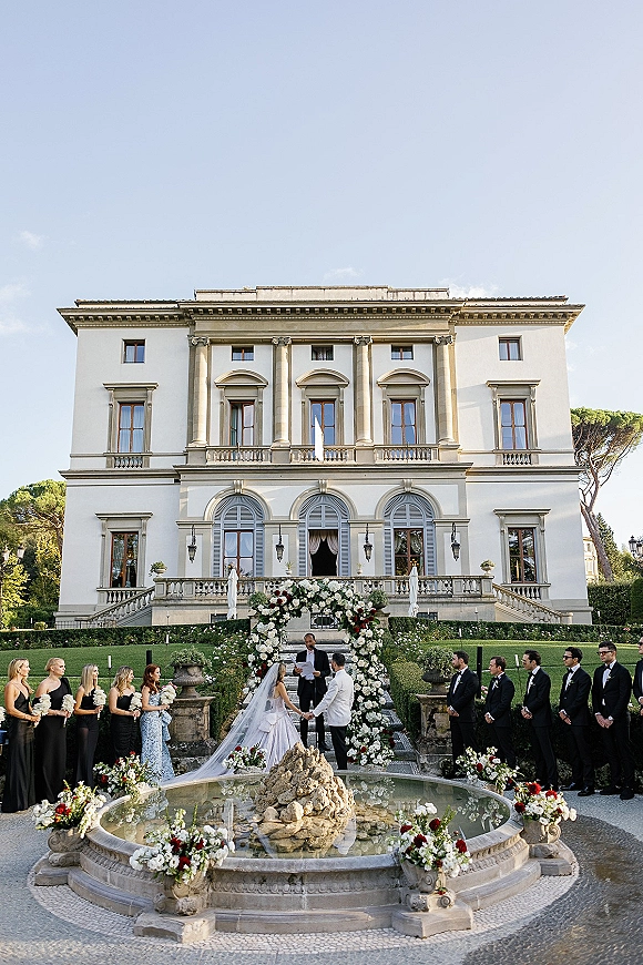 Wedding ceremony with bride and groom under a floral arch, guests lined along aisle flowers by a fountain at a villa garden lawn