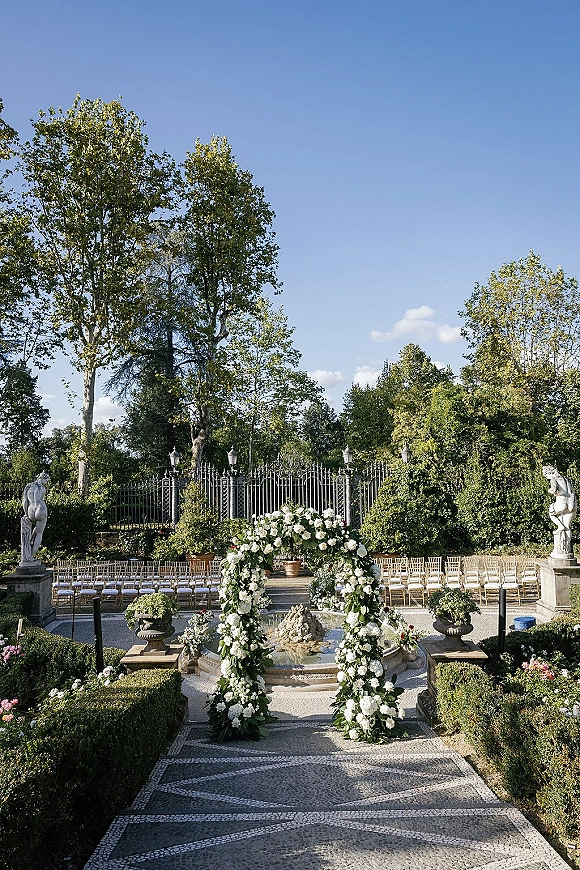Outdoor ceremony setup with a floral arch framed in white flowers and greenery, aisle seating facing a fountain in a formal garden courtyard