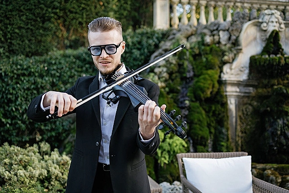 Wedding musician in a black tuxedo playing violin in sunglasses beside a stone fountain, framed by garden greenery and outdoor seating