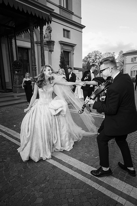 Wedding reception moment as the bride in a strapless ball gown laughs with a violinist under a mansion portico, lantern overhead