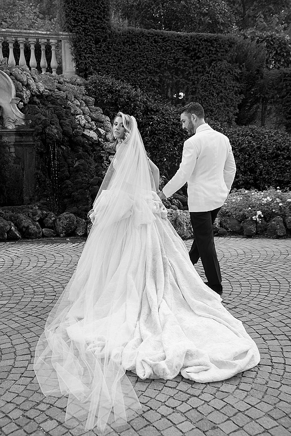 Couple portrait of bride and groom walking away hand in hand, bride glancing over shoulder as her veil and long train sweep a cobblestone courtyard by a stone fountain