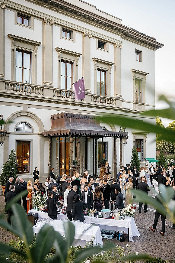 Wedding cocktail hour with black-tie guests mingling by a white-linen cocktail bar, floral centerpieces and lanterns in a grand courtyard facade backdrop