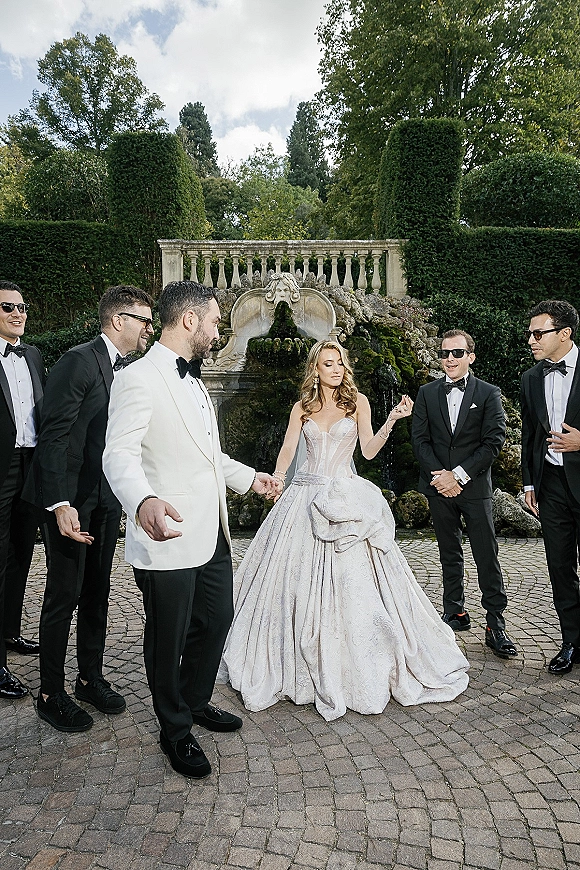 Wedding party portrait of a bride with groomsmen in tuxedos and sunglasses beside a stone fountain in a cobblestone garden courtyard