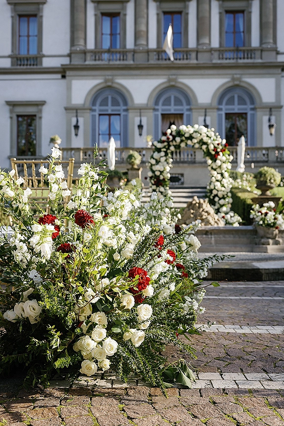 Ceremony setup with a floral arch and gold chiavari chairs, framed by a stone courtyard fountain and villa facade with arched windows.
