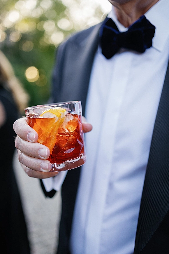 Wedding cocktail in a rocks glass with ice and orange peel garnish, held by a groom in tuxedo amid greenery and bokeh lights