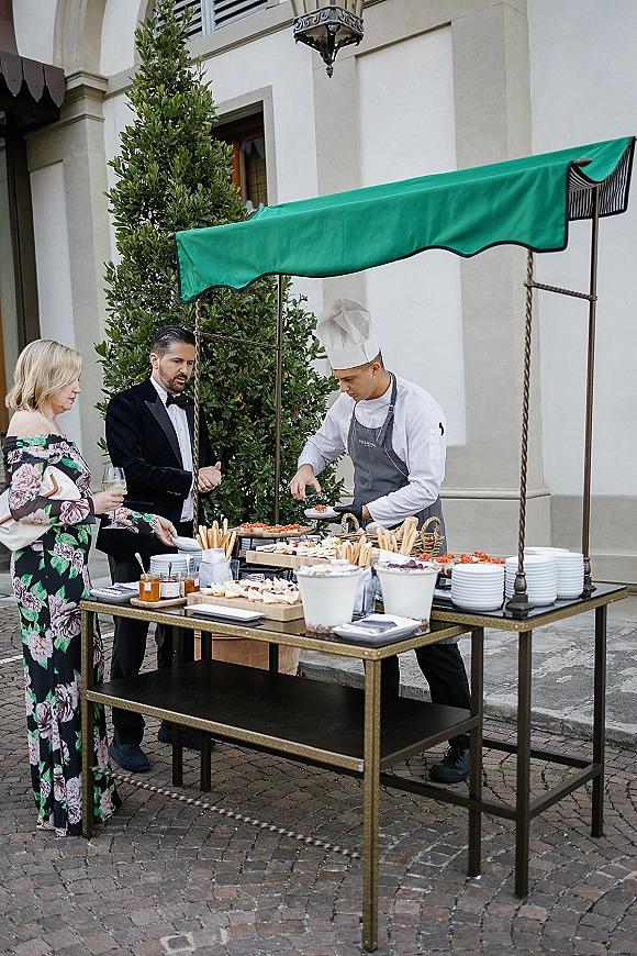 Wedding cocktail hour featuring a wedding catering cart with chef in hat serving hors d'oeuvres under a green canopy in a stone courtyard