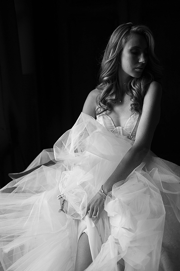 Bridal portrait of a bride in a strapless wedding dress with a tulle skirt, jewelry and rings, looking down in moody window light