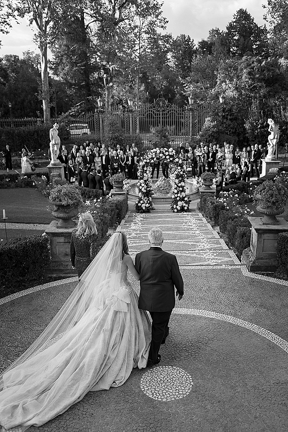 Wedding processional as bride walks down aisle with her father, long cathedral veil and gown train on a stone mosaic garden walkway