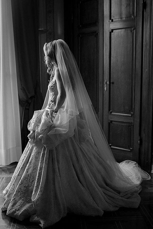 Bridal portrait in black and white of a bride by window, wearing a strapless lace gown with cathedral veil and long train in soft light
