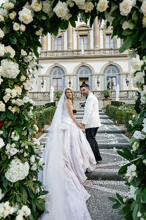 Couple portrait of bride and groom holding hands, looking back on stone steps under a white rose floral arch at a grand estate facade