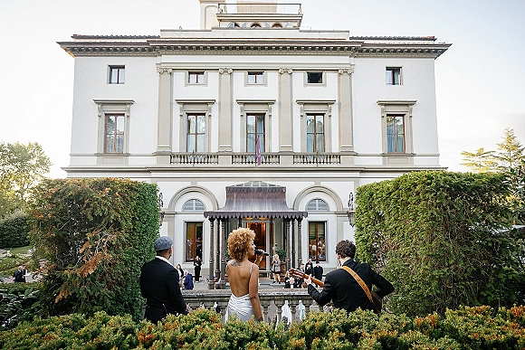 Wedding live music with a singer at a microphone and guitarist performing on a lawn outside a classic villa entrance as guests watch
