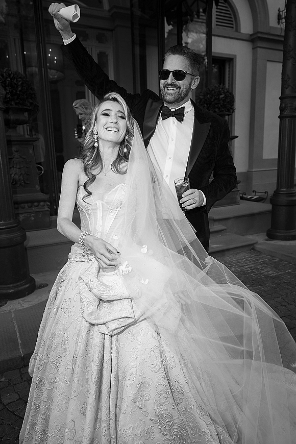 Wedding exit moment as bride and groom laugh walking down steps amid flower petals, groom in sunglasses by columned entrance