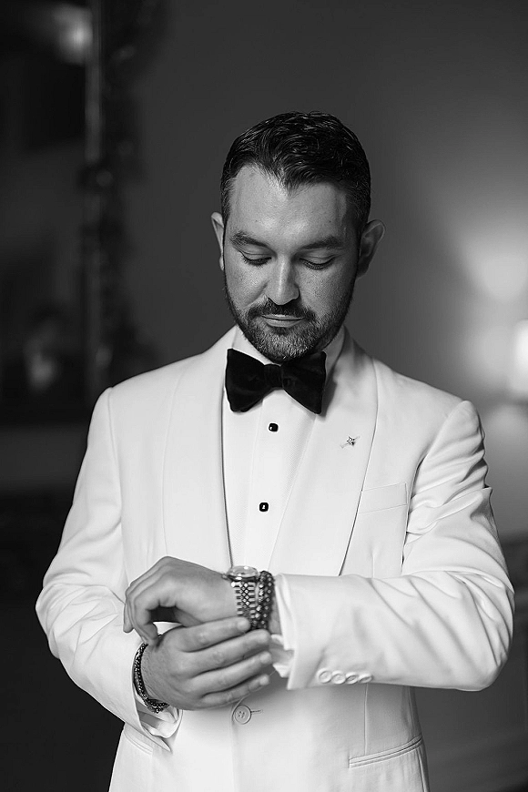 Groom portrait in a white tuxedo jacket and black bow tie adjusting his wristwatch in an indoor hallway with blurred lights