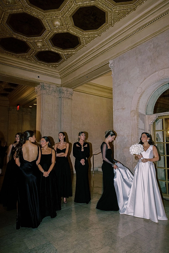 Bridesmaids with bride in black dresses admire the bride holding a round white bouquet under an ornate ceiling by arched doors