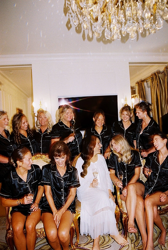 Bridesmaids getting ready in matching satin pajamas raising champagne flutes, smiling together under a chandelier in a living room