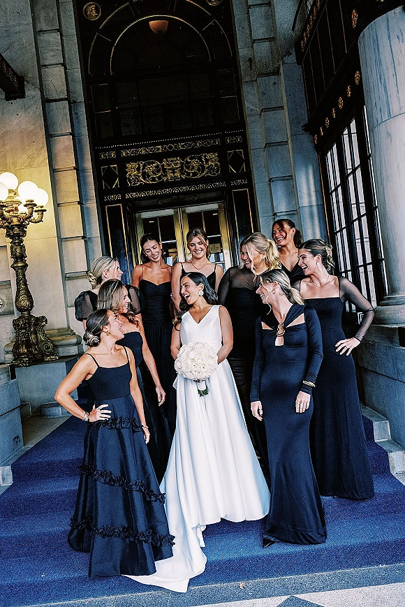 Bride with bridesmaids in black dresses gathered on stone steps, bride in white gown holding a white bouquet at ornate entrance