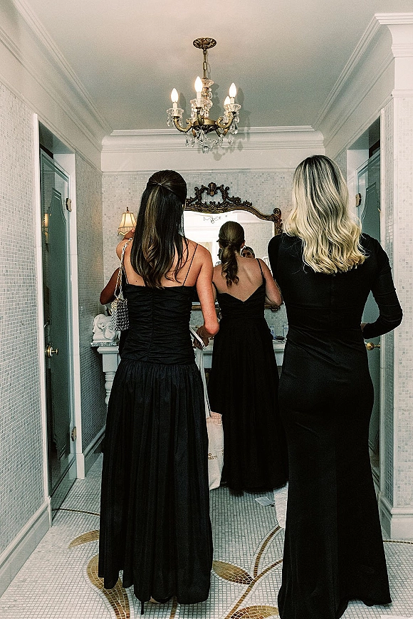 Bridesmaid getting ready with bridesmaids in black dresses, one fixing her hair by an ornate mirror in a mosaic-tiled bathroom under a chandelier