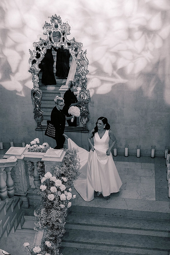 Bride portrait on stairs in a satin wedding dress with veil, holding her train beside roses, baby's breath, and pillar candles on stone steps