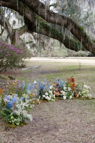 Ceremony altar decor with a clear acrylic lectern and ground floral arrangements beneath oak trees draped in hanging moss on a lawn