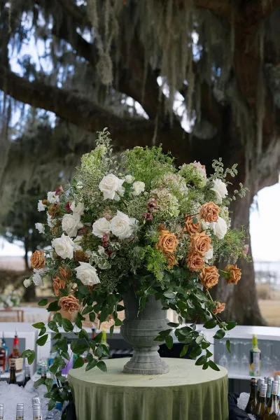 Floral centerpiece with white and tan roses and greenery overflowing from an urn vase on a green tablecloth at an outdoor bar setup under an oak tree with hanging moss