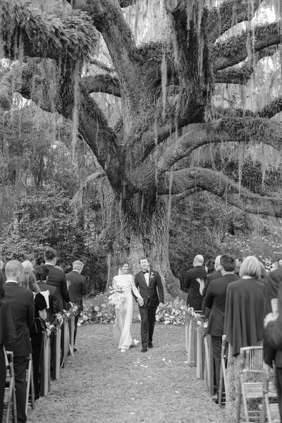Wedding recessional with bride and groom walking aisle, she holds a cascading bouquet as petals fly under an oak tree with hanging moss.