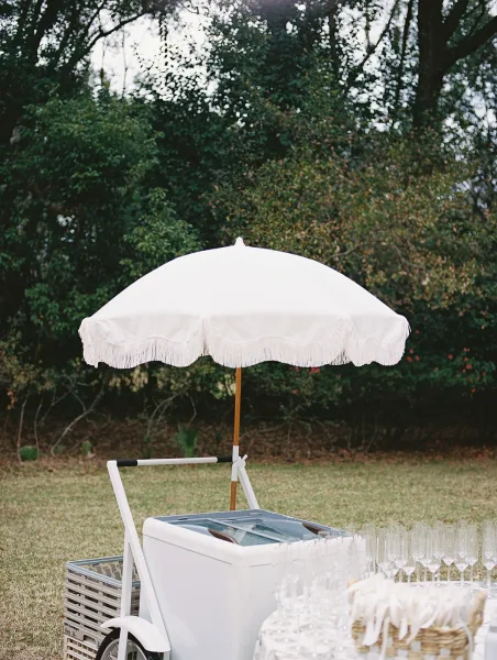 Champagne cart setup with wedding champagne cart under a white fringe umbrella, flutes and rattan basket on a grassy garden lawn