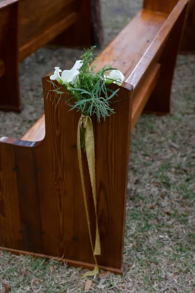 Ceremony pew florals with white calla lilies and greenery tied with ribbon to wooden pew ends beside a simple grass-and-dirt aisle